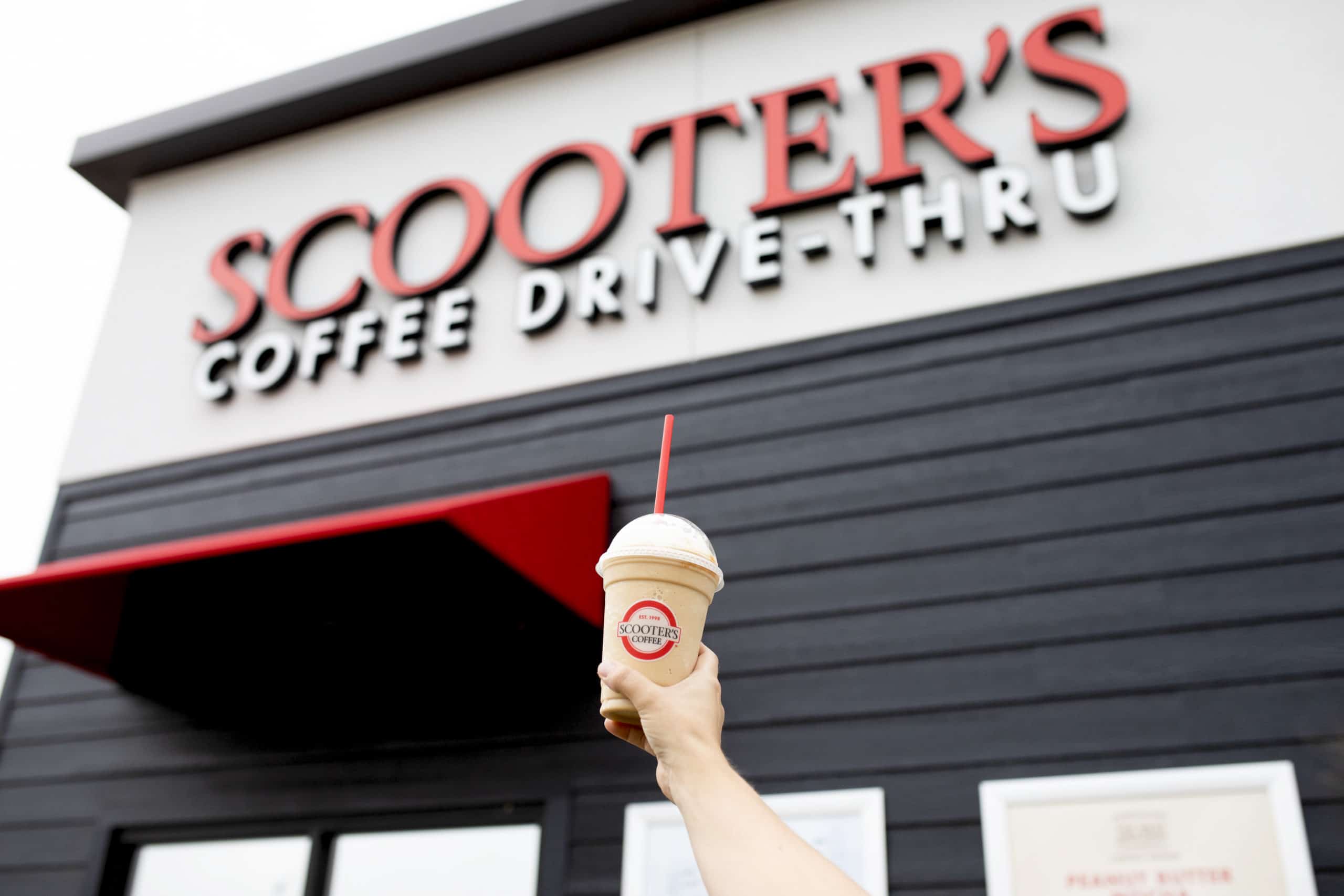 A person triumphantly holding a blended coffee drink in front of a Scooter's Coffee location.