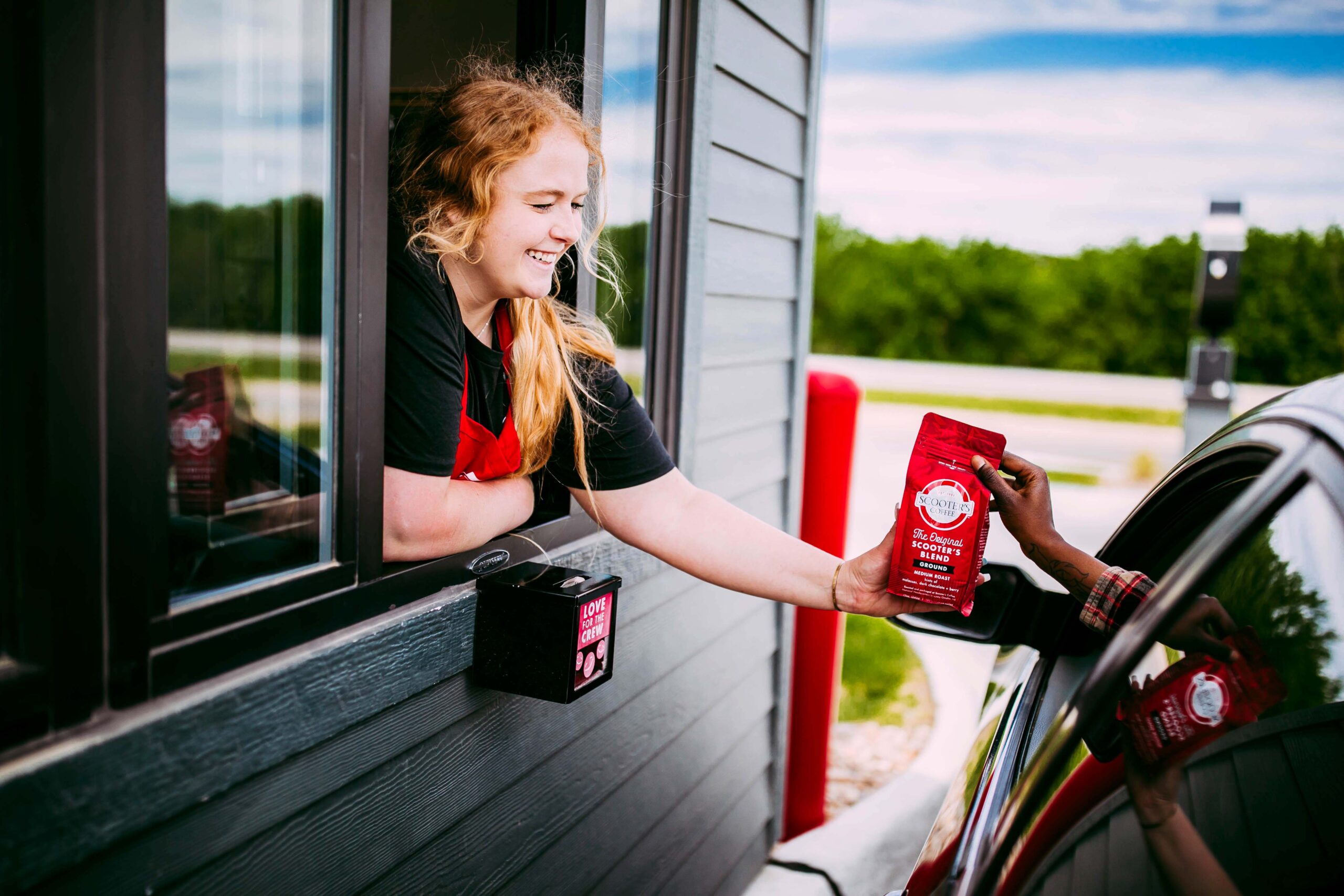 Red headed barista handing coffee bag to customer in car whose hand is only available