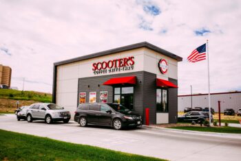 Outside view of a Scooter's Coffee kiosk on a cloudy day with cars in the drive thru and the american flag in the background