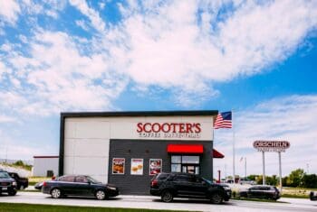 Scooters coffee kiosk with american flag, cars in the drive through and big blue skies in the background